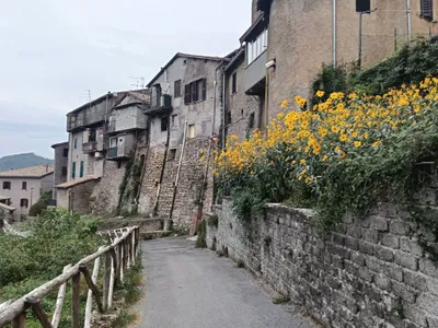Via Santa Maria: la stradina con la staccionata in legno, le vecchie case affacciate sul pendio e il muro in pietra coperto di fiori gialli.