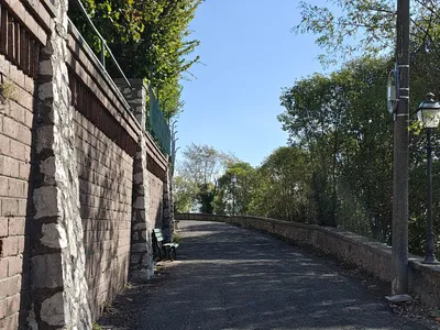 La strada che gira attorno al colle, sotto il muro in mattoni e pietra della chiesa, con una panchina verde e i lampioni sul lato della valle.