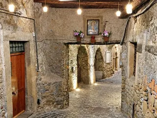 Il passaggio coperto di sera: il soffitto di travi in legno, le lampade accese e un'edicola sacra con i fiori sopra l'arco.