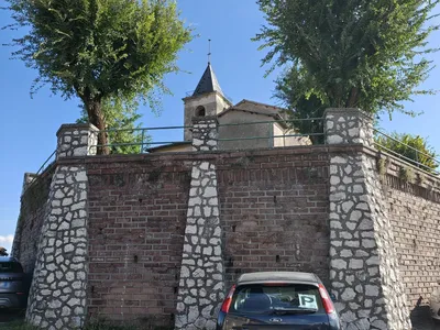 Il muro in mattoni con i contrafforti in pietra bianca che sostiene il prato della chiesa; sopra, il campanile a punta.