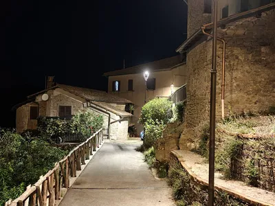 La stradina di sera, illuminata dai lampioni: a sinistra la staccionata in legno affacciata sul verde, a destra il muretto e le vecchie case in pietra.