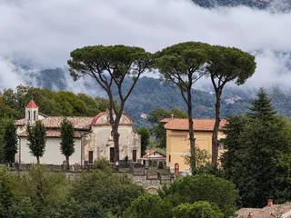 Il santuario visto da lontano, sotto tre grandi pini a ombrello, con le nuvole basse sui monti alle spalle.