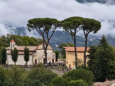Il Santuario della Madonna di Compigliano visto da lontano, sotto tre grandi pini a ombrello, con le nuvole basse sui monti alle spalle.