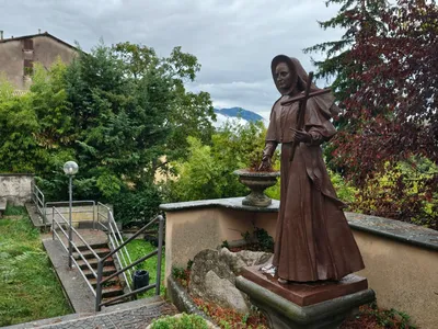 Una statua in bronzo di una religiosa con la croce in mano, su un terrazzino con una lapide ai suoi piedi, tra gli alberi e con i monti sullo sfondo.