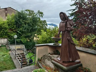 Una statua in bronzo di una religiosa con la croce in mano, su un terrazzino con una lapide ai suoi piedi, tra gli alberi e con i monti sullo sfondo.