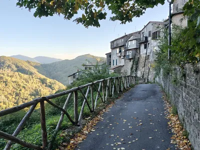 La stradina con la staccionata in legno in una mattina d'autunno: le foglie gialle sull'asfalto, le vecchie case affacciate sul pendio e le montagne illuminate dal sole.