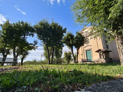 Il prato alberato accanto alla chiesa, con le panchine, i lampioni e il pozzo in pietra con la tettoia.