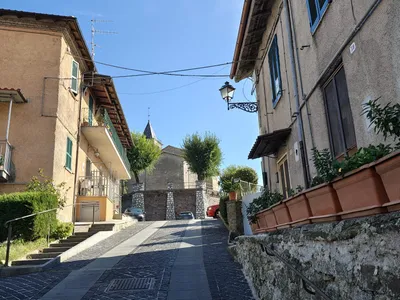 Via del Monte sale tra le case con le persiane e i vasi di fiori verso il muro con i contrafforti in pietra bianca, sotto il campanile della chiesa.