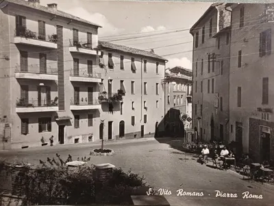 Piazza Roma in una cartolina d'epoca in bianco e nero: i palazzi con i balconi fioriti, l'aiuola con un alberello al centro e, sulla destra, i tavolini all'aperto del bar.