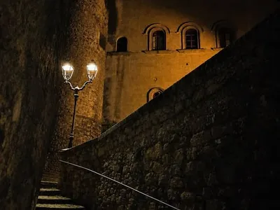 Il castello di notte, visto dalla scalinata in selciato nei pressi della Chiesa di Santa Maria de Arce: la torre rotonda e le finestre ad arco alla luce dei lampioni.