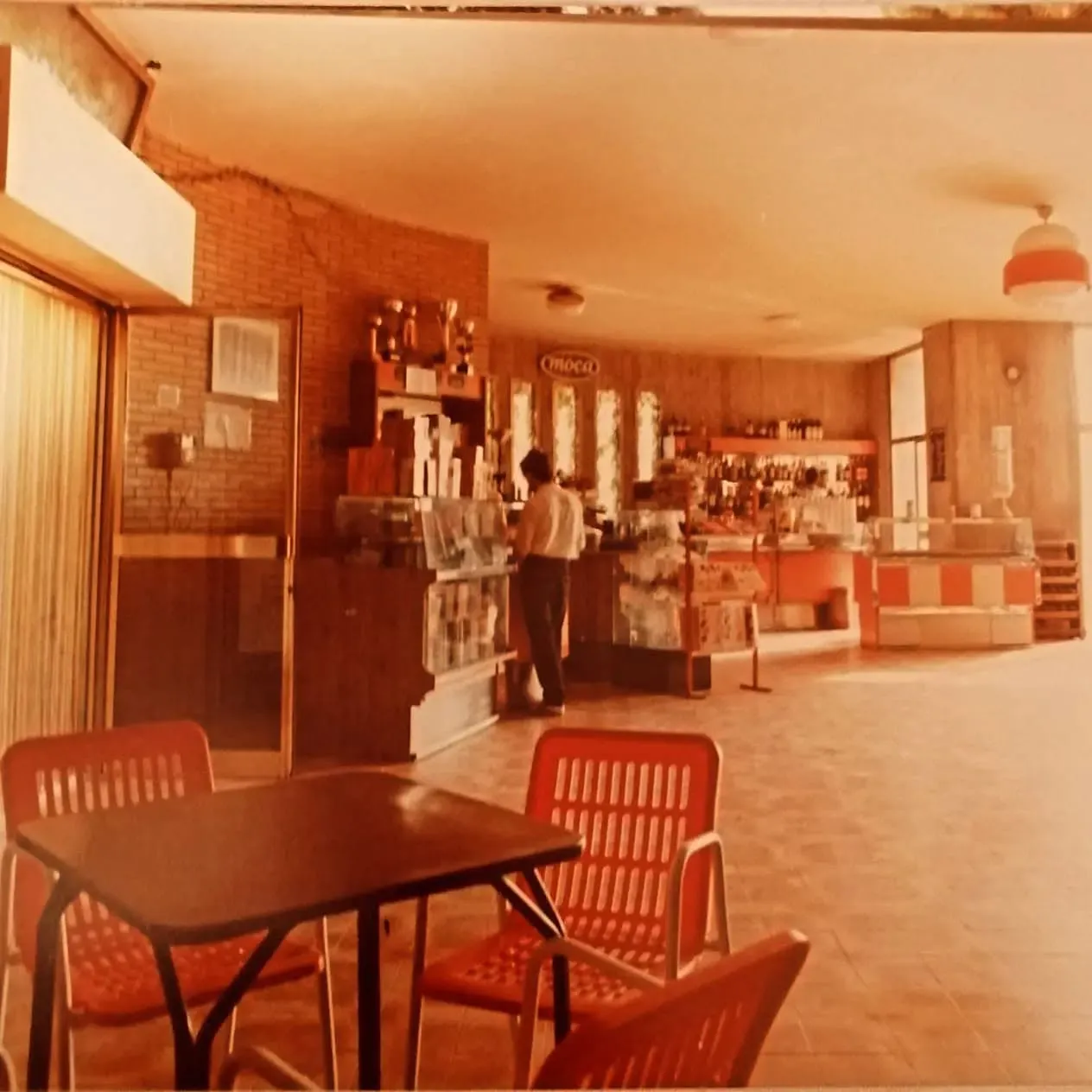 Il bancone e gli scaffali del bar, con i tavolini dalle sedute rosse in primo piano.