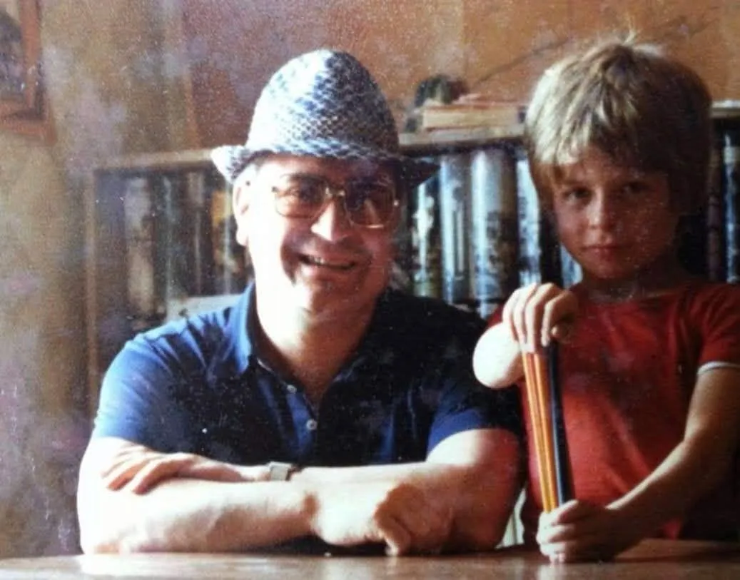 Don Marcello, con il cappello estivo, seduto al tavolo accanto al piccolo Giammarco, che stringe un fascio di matite colorate.