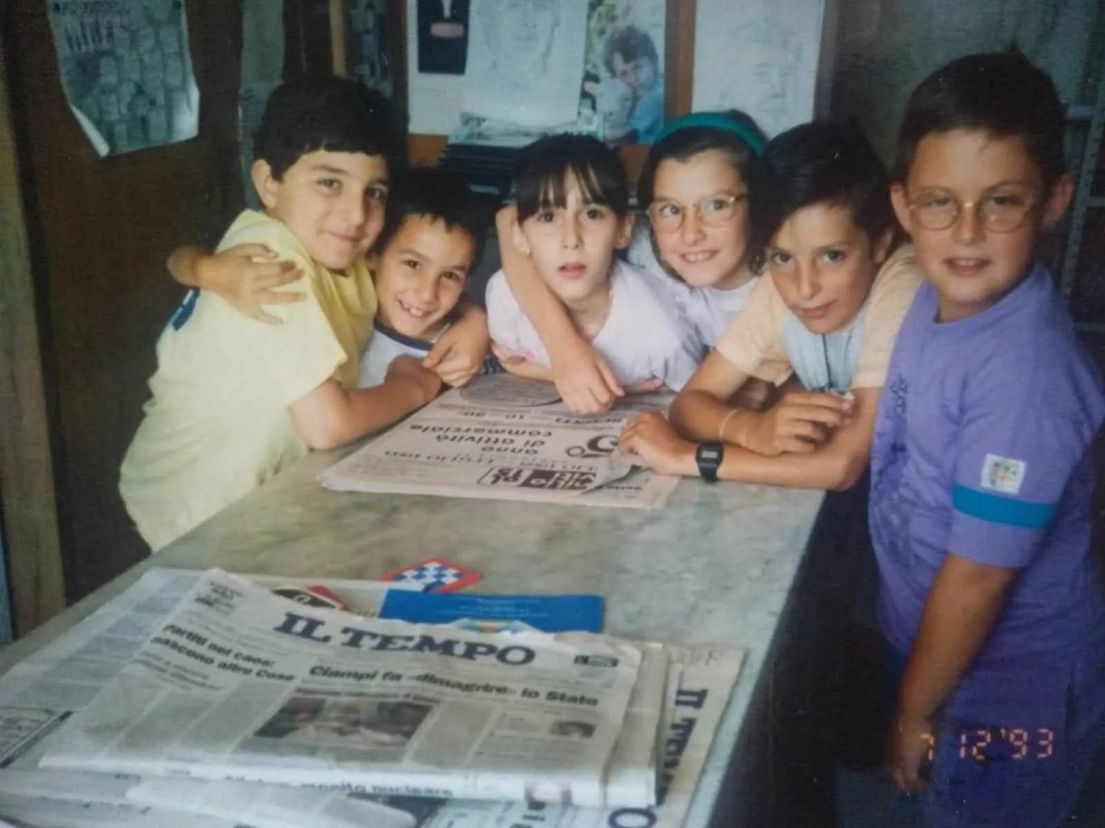 Un gruppo di bambini nello studio e biblioteca di Don Marcello, abbracciati attorno al tavolo di marmo coperto di giornali; la data stampata sulla foto è del 1993.